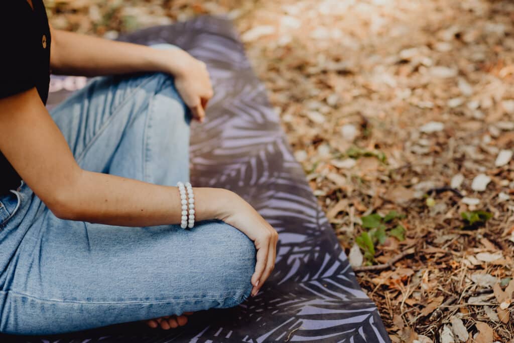 Méditation en pleine nature : calme et bien-être. Femme méditant en tailleur sur un tapis de yoga à motifs, portant des jeans et bracelets en pierre, au milieu de feuilles sèches.