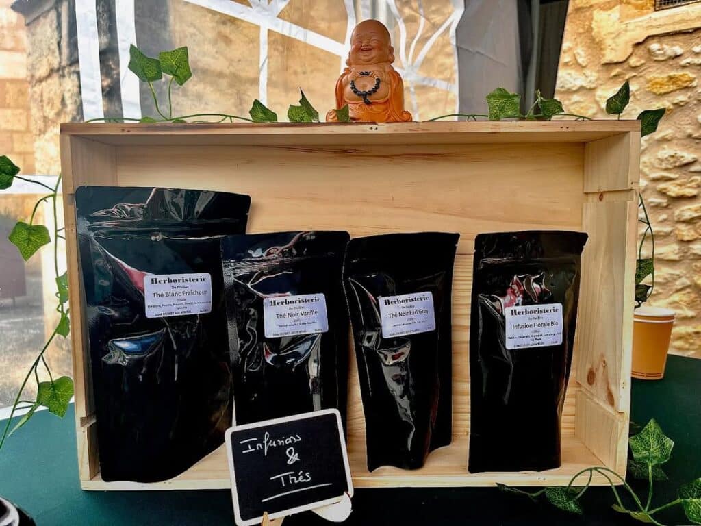 Herboristerie : Thés & Infusions Naturelles et Biologiques Étalage d'herboristerie : caisse en bois, Bouddha souriant, lierre. Quatre sachets de thés et infusions, dont Thé Blanc Fraîcheur, Thé Noir Earl Grey. Panneau : 'Infusions & Thés'.